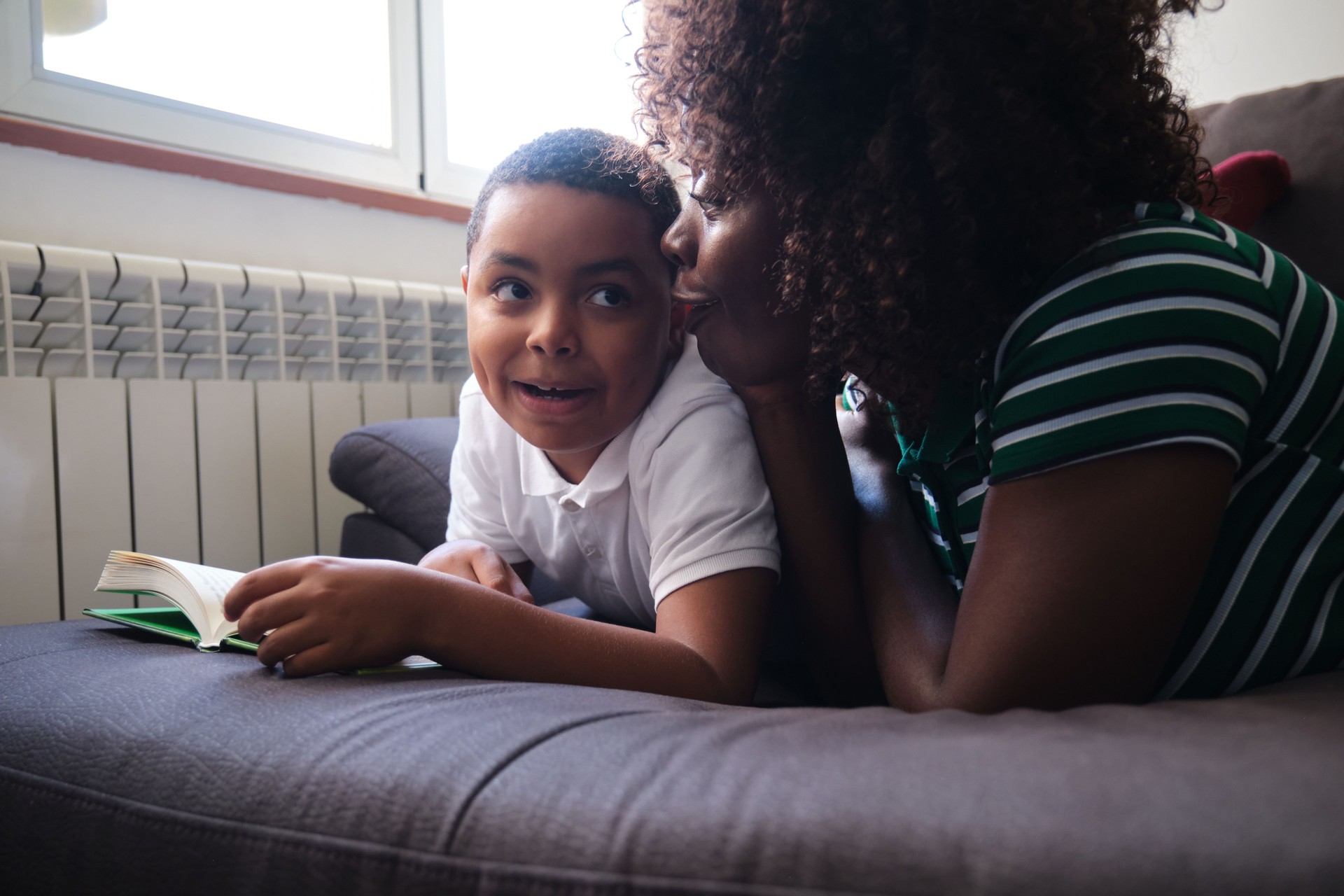 Mother whispering to her son reading a book lying on the sofa, doing funny faces