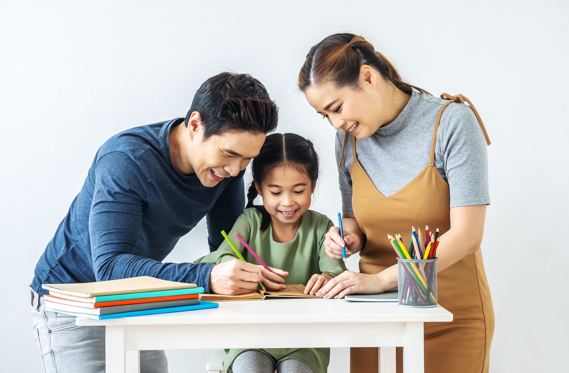 Asian young mother and father with little daughter sit at desk learning and writing in book with pencil making homework at home.Education concept.